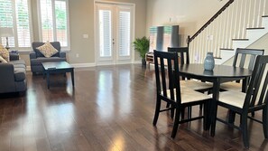 Ein geräumiges Wohnzimmer mit dunklem Holzboden, einem Essbereich mit rundem Tisch und Stühlen sowie einer Treppe mit weißen Geländern.
