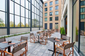 Ein Außenbereich mit Terrasse, Tischen, Stühlen, großen Fenstern und einem Gebäude im Hintergrund.
