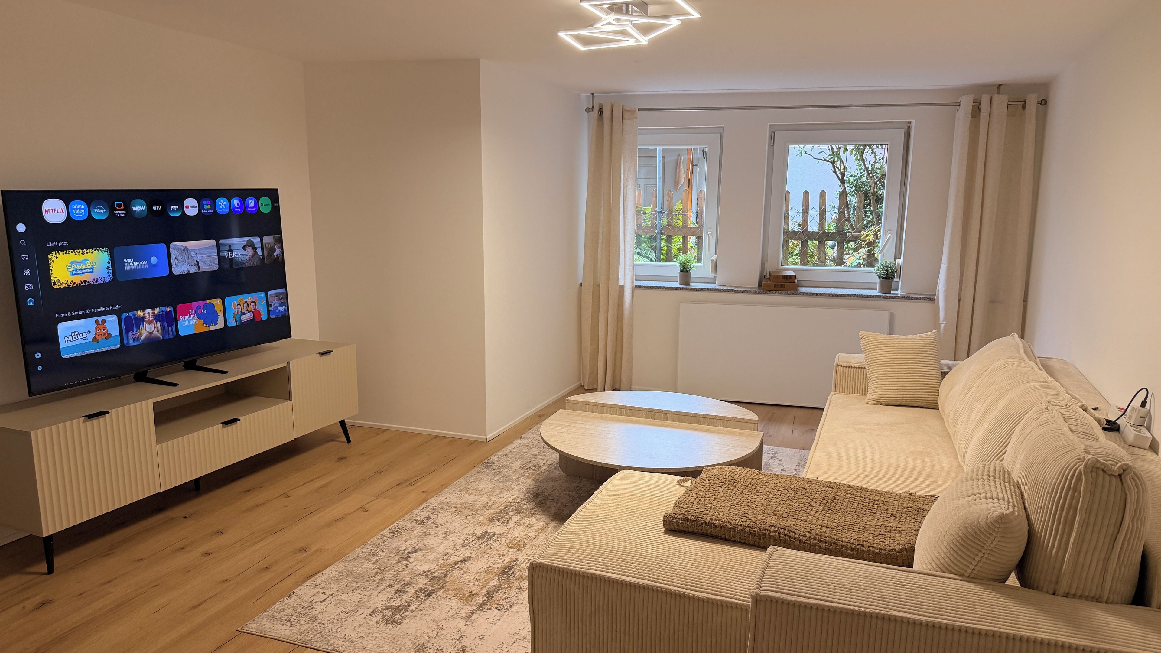 A modern living room with a flat-screen TV, a sofa, a coffee table, and a window with curtains.