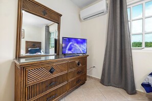 A room with a wooden dresser, a flat-screen TV, an air conditioning unit, and a window with curtains. - Palma Real 29 - your vacation Condo in Palm Beach (Palma Real)