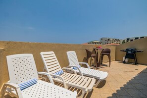 Terraço com cadeiras de plástico brancas, uma pequena mesa e uma churrasqueira.