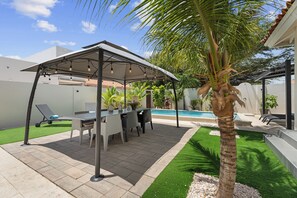 A covered outdoor seating area with a pool, artificial grass, and palm trees. - Modern Three Bedroom Villa in Noord (Boegoeroei)