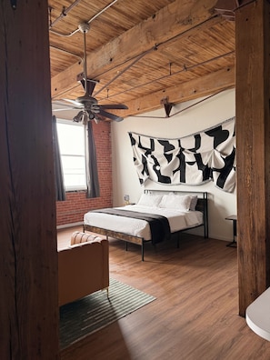 A bedroom with a bed, a nightstand, a chair, a ceiling fan, and a brick wall. - 420-Friendly King suite Loft Walk to Detroit Riverfront FREE parking (Detroit)