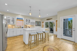 A modern kitchen with white cabinets, a central island with bar stools, and a view to the outside. - OIB Waterfront Retreat w/ Dock & Golf Cart 5BR (Ocean Isle Beach)
