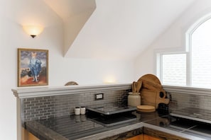 A kitchen with a brick backsplash, a wooden stool, a painting, and a window with blinds. - Charming Farmhouse Near Calgary & Spruce Meadows (East)