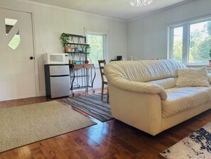 A living room with a beige sofa, a wooden floor, a kitchen area with a refrigerator and microwave, and a window with natural light.
