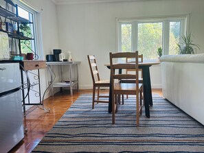 A compact kitchen with a refrigerator, a wooden dining table, and chairs. There is a window providing natural light and a view of greenery outside.