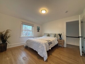 Quarto com cama grande, mesa de cabeceira com planta, janela com vista e porta que dá para outro cômodo.