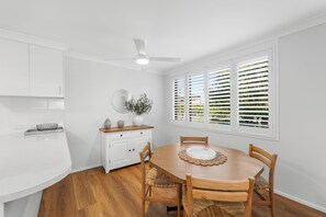 Een moderne keuken met witte kastjes, een houten eettafel met vier stoelen en een groot raam met jaloezieën.