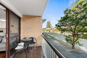 Un balcon avec un mur en briques, une table, deux chaises et une vue sur une rue bordée d’arbres et où circulent des voitures.
