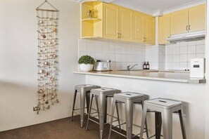 Une cuisine avec des meubles de couleur jaune, un plan de travail blanc, un évier, un grille-pain, une plante et trois tabourets de bar.