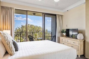 Une chambre d’hôtel comprenant un lit, une télévision, une commode en bois, un balcon avec vue sur les arbres et les bâtiments, et une chaise.