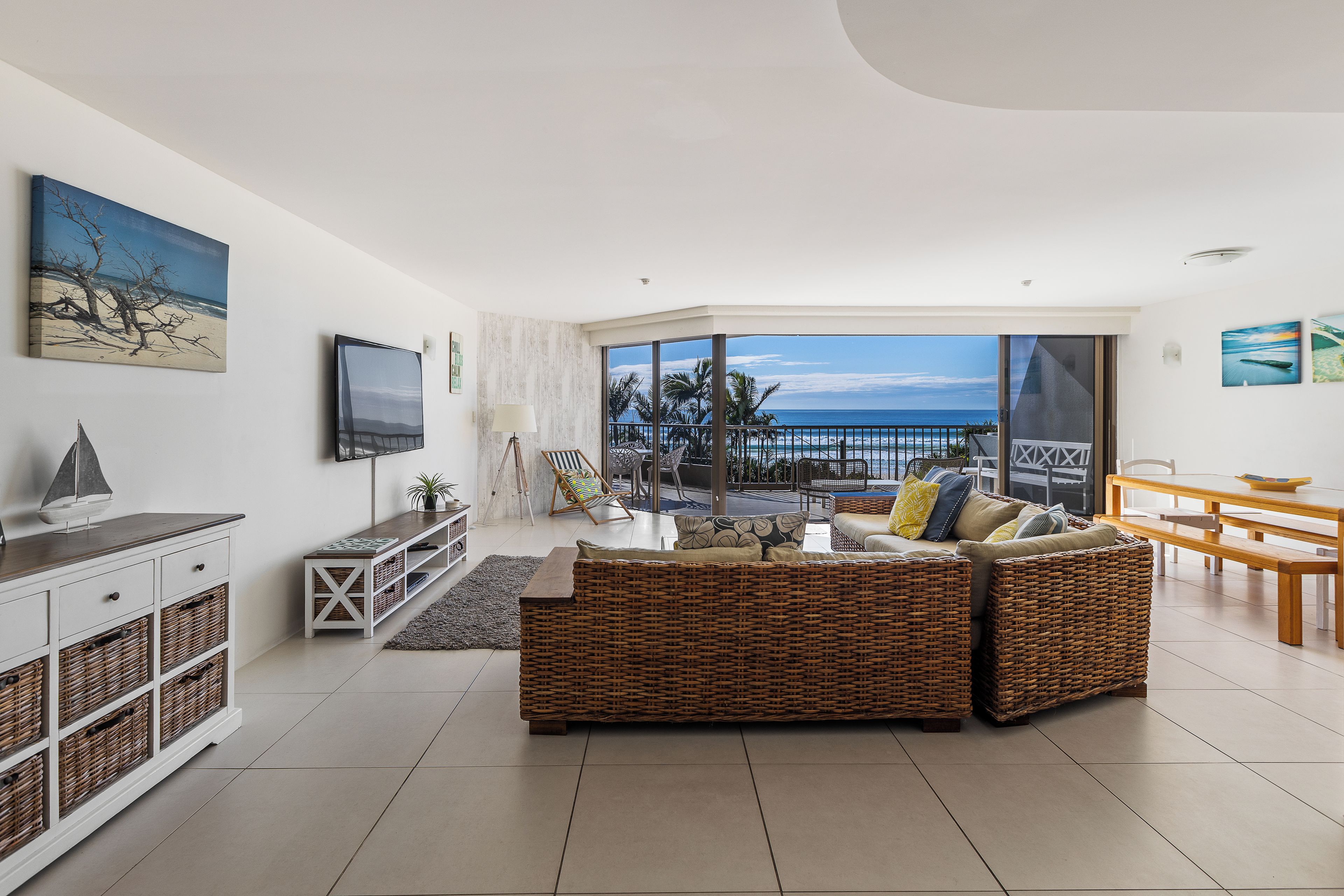 Een moderne woonkamer met een groot raam met uitzicht op een strand, een flatscreen televisie aan de muur en een eethoek met een houten tafel en bankjes.