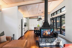 A modern kitchen with white cabinets, a dining area with a table and chairs, and a large fireplace with a stone surround. - Whispering Creek - Luxury Off Grid Escape (Currumbin Valley)