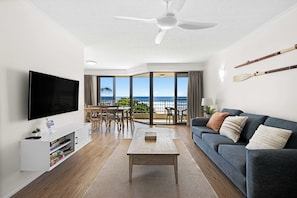 A modern living room with a flat-screen TV, a sofa, a coffee table, and a dining area with a view of the ocean. - Surfside 8 with direct beach access. (Palm Beach)