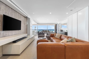 A modern living room with a large flat-screen TV mounted on a wooden wall, a leather sofa, and a dining area with a view of the ocean. - Joy on Jefferson Stunning Palm Beach Escape (Palm Beach)