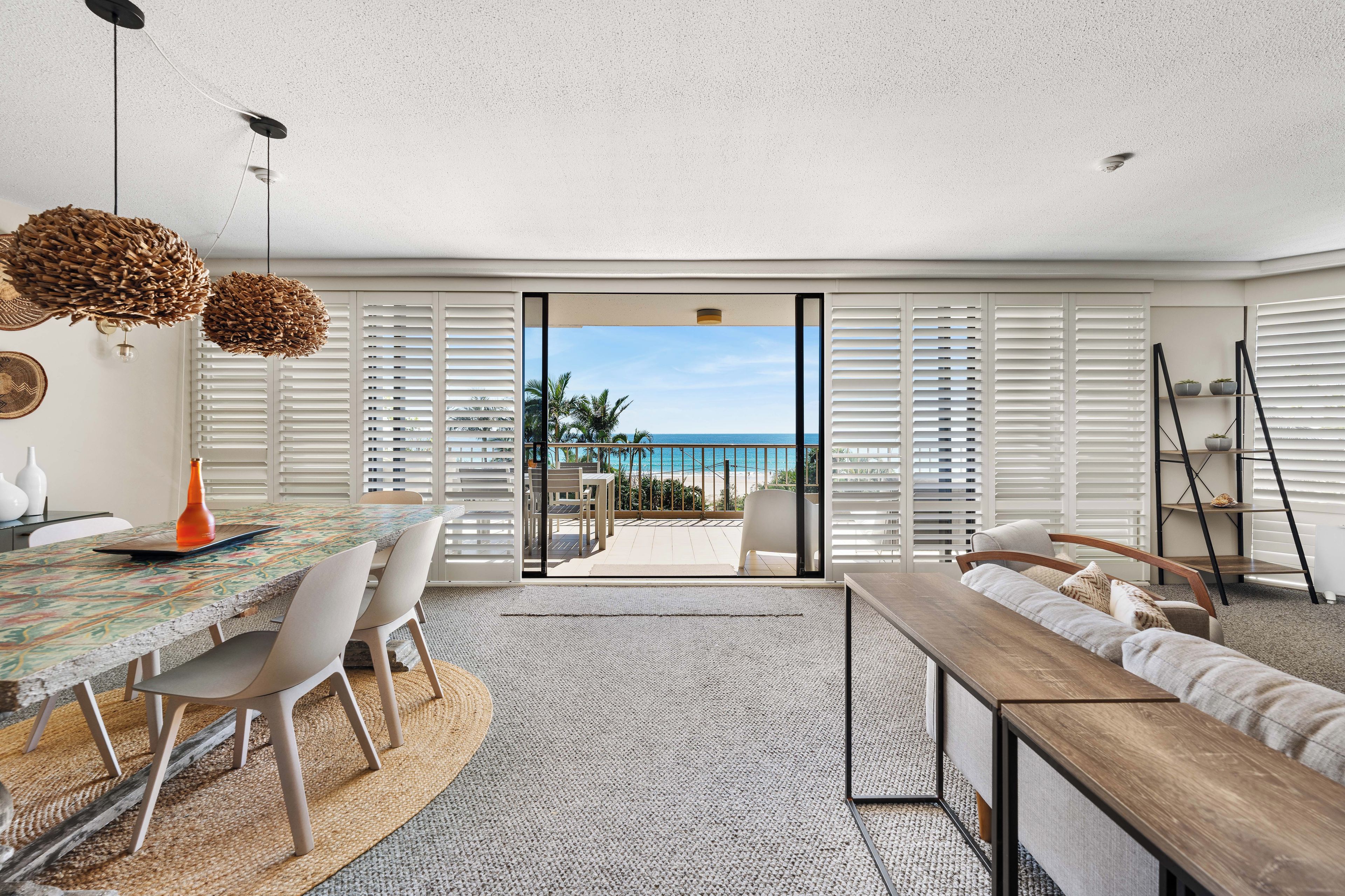 Een moderne woonkamer met een groot raam met uitzicht op een strand, een eethoek met een kleurrijke tafel en een gezellige zithoek met een houten bank.