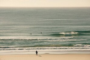 A beach with a person standing on the sand, waves approaching the shore, and a few people in the water. - The Rocks Resort 4C Currumbin Beachfront Apartment (Currumbin)