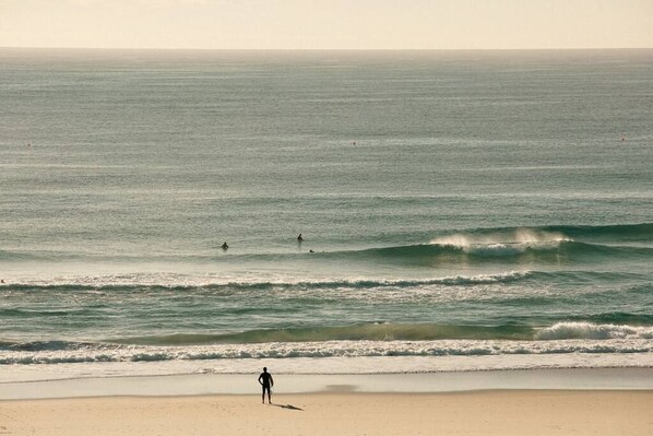 A beach with a person standing on the sand, waves approaching the shore, and a few people in the water. - The Rocks Resort 4C Currumbin Beachfront Apartment (Currumbin)