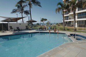 A swimming pool with lounge chairs and umbrellas, surrounded by palm trees and a building with balconies. - Golden Era - Tugun Beachfront (Tugun)