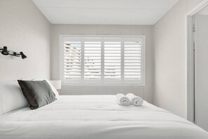 A minimalist bedroom with a bed, a window with blinds, a bedside lamp, and rolled towels. - White Dolphin 7 Beachfront Currumbin Apartment (Currumbin)