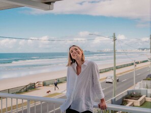 Une personne se tient sur un balcon donnant sur une plage, avec une route et un chemin piétonnier en arrière-plan.