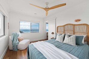 Une chambre avec un grand lit, une vue sur l’océan par la fenêtre, un ventilateur de plafond et une tête de lit en bois.