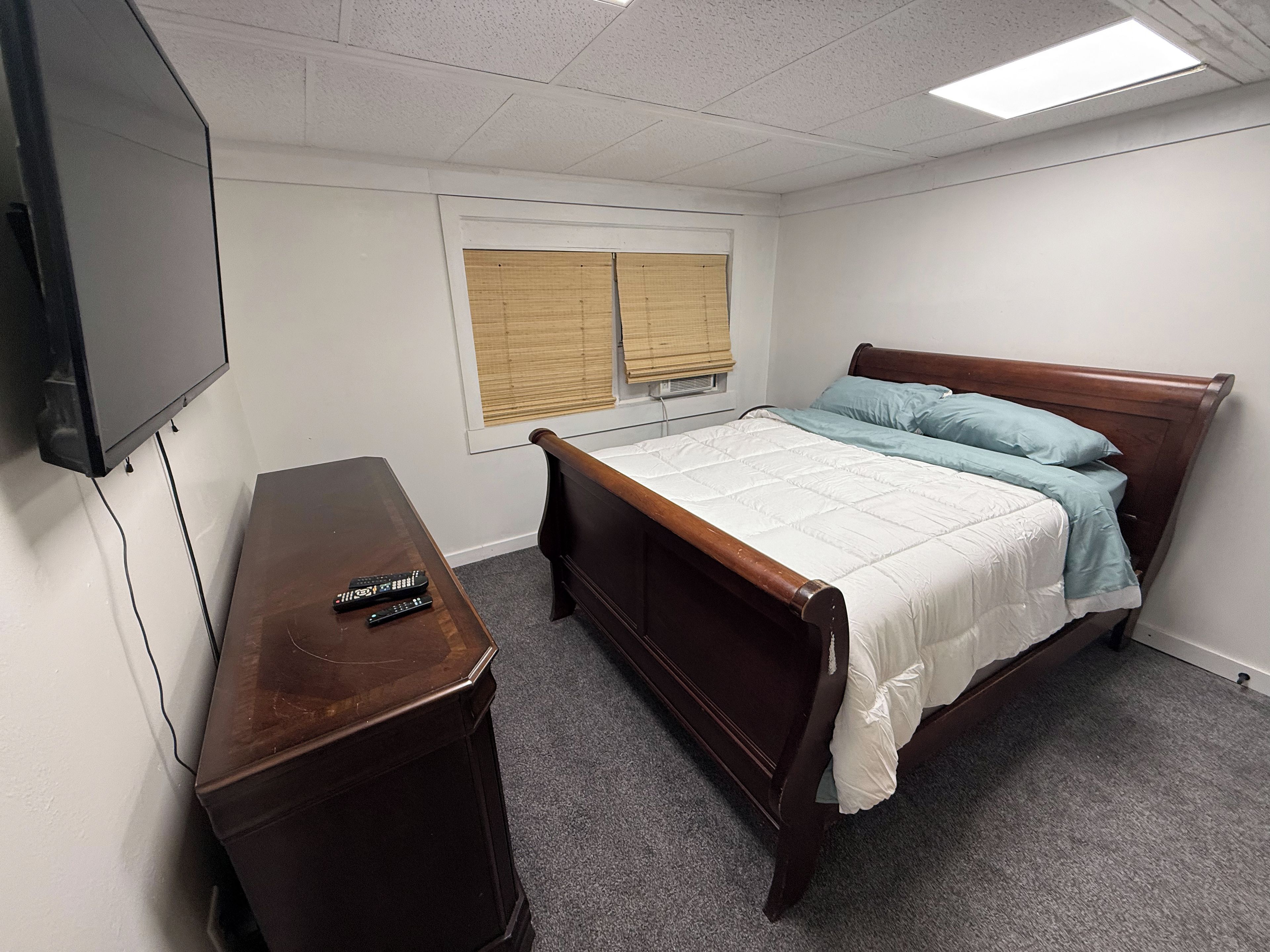 A bedroom with a bed, a wooden nightstand, a television, and a window with blinds.