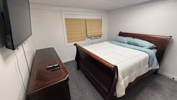 A bedroom with a bed, a wooden nightstand, a television, and a window with blinds.
