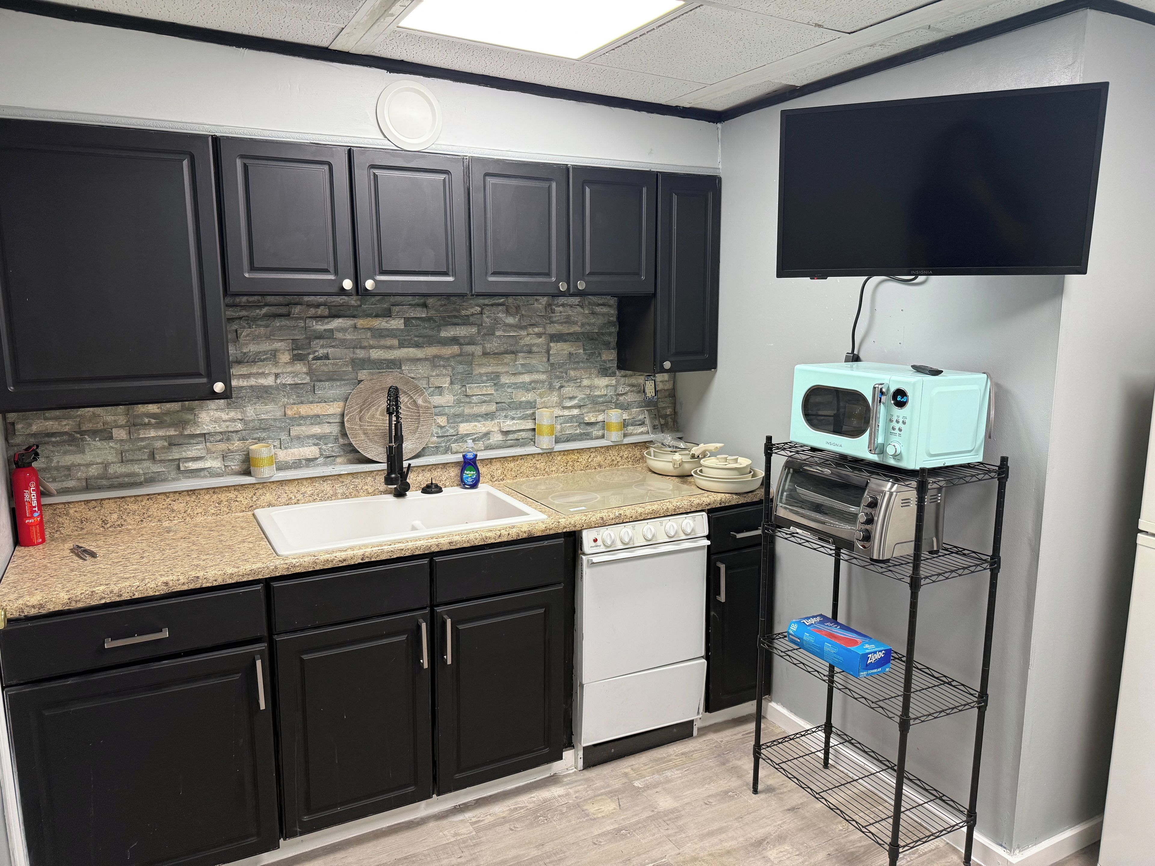 A kitchen with black cabinets, a granite countertop, a built-in dishwasher, a microwave, and a shelf with kitchen items.