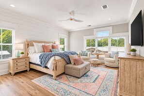Une chambre à coucher comprenant un lit, des tables de chevet, un ventilateur de plafond, une grande fenêtre et un téléviseur fixé au mur.