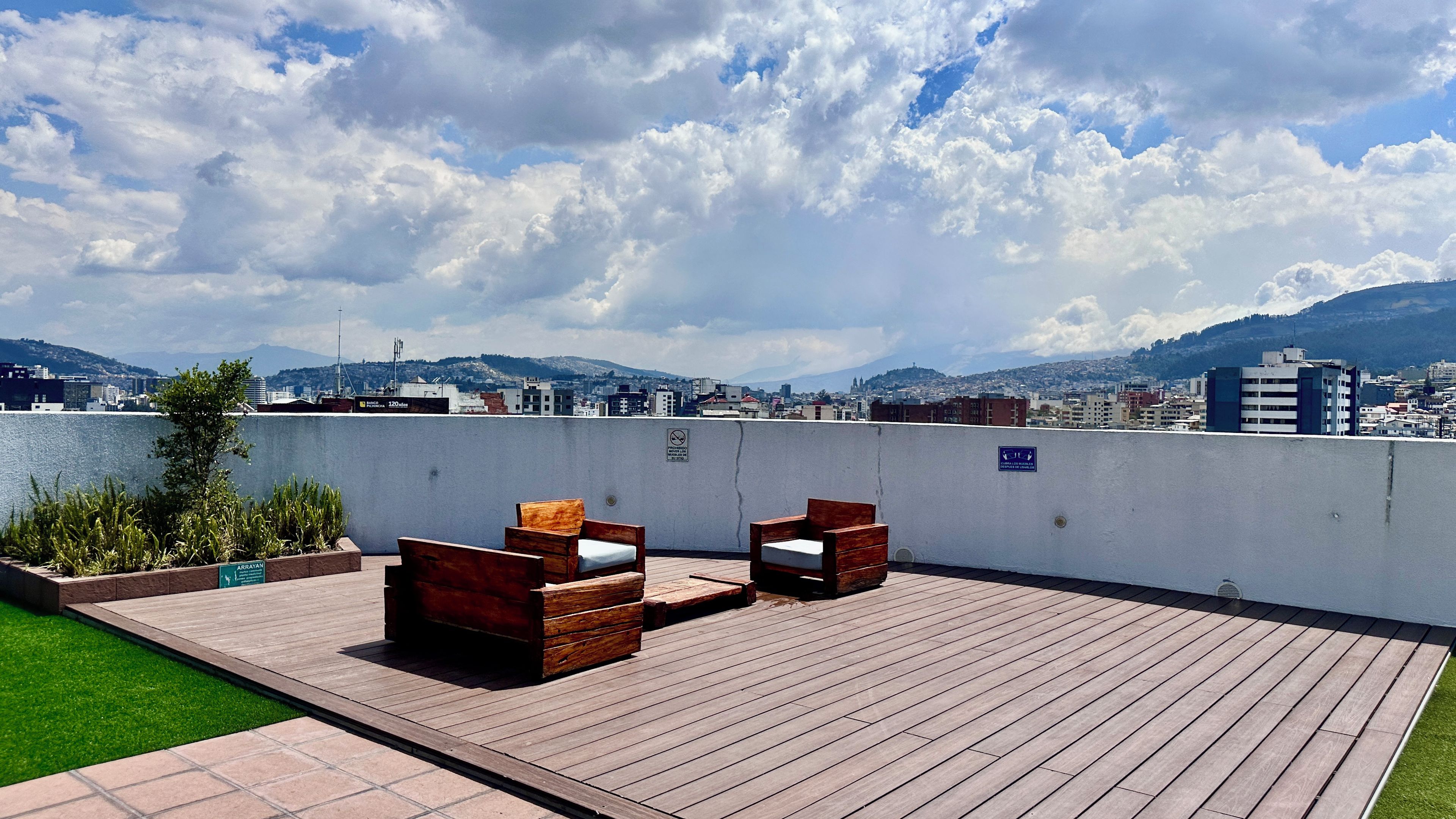 A rooftop terrace with wooden furniture, a view of a cityscape, and a sign indicating a smoking area.