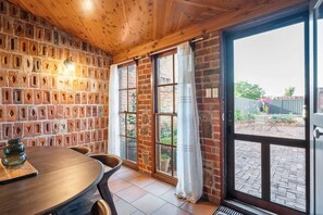 A room with a brick wall, wooden ceiling, a round table with chairs, and a sliding glass door leading to an outdoor area. - Sheiling 1888  A Honey Home (Dubbo)