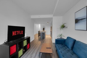 A modern living room with a flat-screen TV displaying 'NETFLIX', a blue sofa, a wooden coffee table, and a dining area in the background. - The Orient - Spectacular CN Tower Views (Toronto)