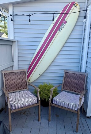Deux fauteuils en osier avec des coussins à rayures sur une terrasse en bois, une planche de surf appuyée contre un mur de maison et des guirlandes lumineuses suspendues au-dessus.