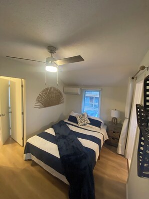 Une chambre à coucher comprenant un lit, un ventilateur de plafond, un appareil de climatisation, une table de chevet et une fenêtre avec des stores.