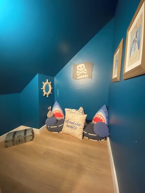 Une pièce aux murs bleus, ornée d’un panneau représentant un crâne et des tibias croisés, de coussins à motifs de requins, d’un coffre au trésor et de tableaux encadrés.