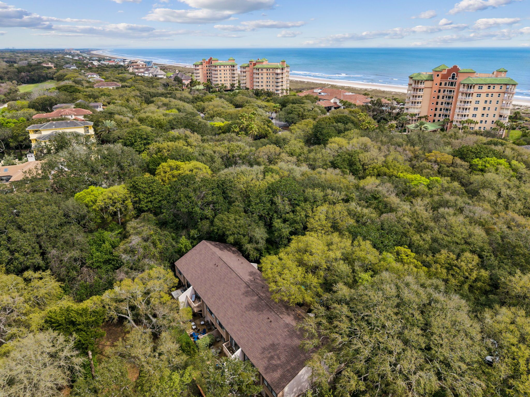 A coastal area with multiple buildings, dense greenery, and a beach in the background.