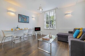 A modern living room with a glass coffee table, a grey sofa, a dining area with chairs, a flat-screen TV, and a large window with blinds. - Contemporary 3BR | Near City Centre | FREE Parking (Reading)