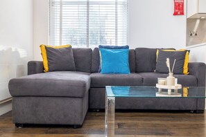 A modern living room with a grey sectional sofa, a glass coffee table, and a window with blinds. - Contemporary 3BR | Near City Centre | FREE Parking (Reading)