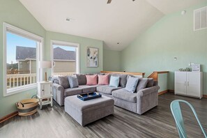 A living room with a sectional sofa, a small ottoman, a white cabinet, and a window with a view of a house. - Beach Please OBX | 1,550 ft to the beach | Private Pool, Hot Tub, and Dog Friendly (Corolla)