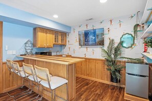 A kitchen with wooden cabinets, a bar area with stools, a refrigerator, and a wall with a poster. - Beach Please OBX | 1,550 ft to the beach | Private Pool, Hot Tub, and Dog Friendly (Corolla)