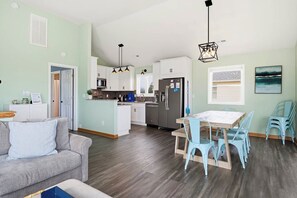 Eine moderne Küche mit weißen Schränken, ein Essbereich mit einem Holztisch und blauen Stühlen sowie ein graues Sofa mit einem hellblauen Kissen.