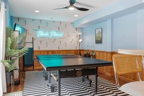 A pool table with a neon sign, a ceiling fan, and a wall with a beach-themed pattern. - Beach Please OBX | 1,550 ft to the beach | Private Pool, Hot Tub, and Dog Friendly (Corolla)