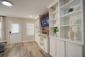 Ein Wohnzimmer mit flachbildfernseher an der Wand, Holzboden, einem grauen Sofa und eingebauten Regalen mit Dekorationselementen.
