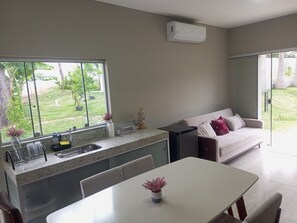 A modern kitchen with a large window, a dining area with a white table, and a living area with a sofa and a sliding glass door.
