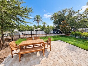 An outdoor patio with a wooden table and chairs, surrounded by a black metal fence, and a well-maintained lawn with a golf flag. - Selah on 30A | Golf Cart, Pool & Hot Tub, Mins to the Beach (Santa Rosa Beach)