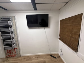A room with a flat-screen TV mounted on the wall, a wooden floor, and a window with blinds. - The Flight Crew Quarters (College Park)