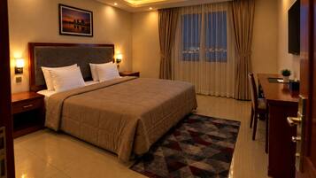 A hotel room with a large bed, a desk with a chair, a nightstand, a lamp, a painting on the wall, and a window with curtains.