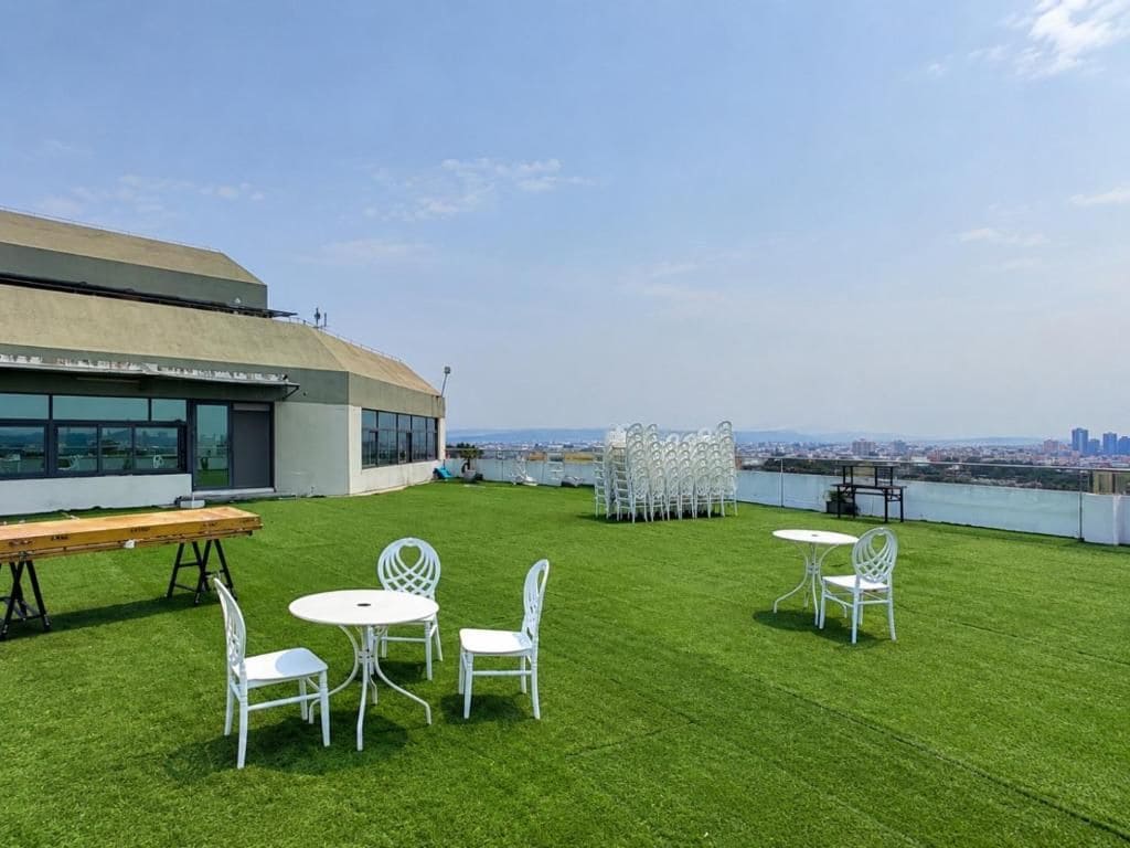 Une terrasse sur le toit offrant une vue sur les toits de la ville, avec un espace repas comprenant des chaises et des tables blanches, ainsi qu’un banc en bois.